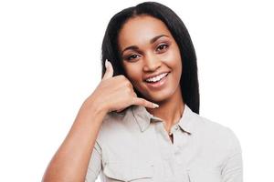 llámame sonriente joven africana gesticulando con el teléfono móvil cerca de su cara y sonriendo mientras está de pie contra el fondo blanco foto