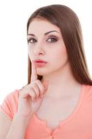 Be quiet Beautiful young woman looking at camera and holding finger on lips while standing isolated on white photo