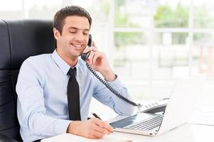 Making some notes. Cheerful young man in formalwear making notes in his note pad and talking on the telephone while sitting at his working place photo