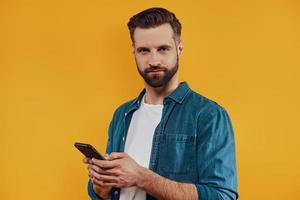 Good looking young man in casual clothing using smart phone and looking at camera while standing against yellow background photo