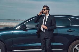 el tiempo es dinero. apuesto joven hombre de negocios hablando en su teléfono inteligente y sonriendo mientras está de pie cerca de su coche al aire libre foto