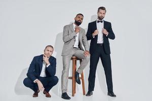 Three handsome young men in suits and bowties looking at camera while standing against gray background photo