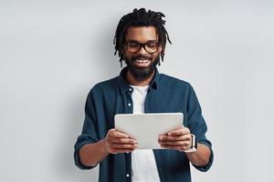 Busy young African man in eyewear using digital tablet and smiling while standing against grey background photo