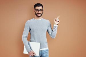 Charming young man in casual wear looking at camera and pointing copy space while standing against brown background photo