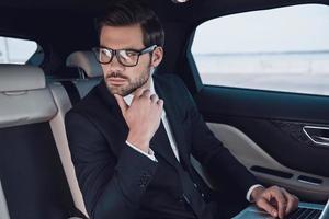 Thinking about business. Thoughtful young man in full suit working using laptop and adjusting his eyewear while sitting in the car photo