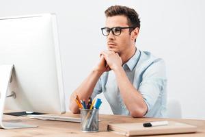 buscando nuevas ideas. hombre guapo joven pensativo con anteojos trabajando en la computadora y manteniendo la mano en la barbilla mientras está sentado en su lugar de trabajo foto