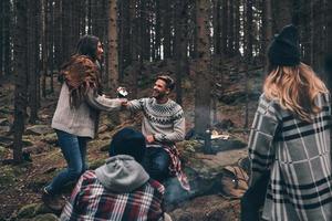 More laugh with friends. Group of happy young people standing around the campfire while hiking in the woods photo