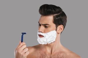 Bad razor Confused young man with foam all over his face looking at razor while standing against grey background photo