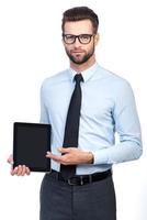 Copy space on his tablet. Confident young handsome man in shirt and tie holding digital tablet and pointing it with smile while standing against white background photo