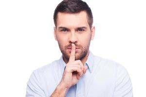 Keep my secret  Serious young man in shirt holding finger on lips and looking at camera while standing against white background photo