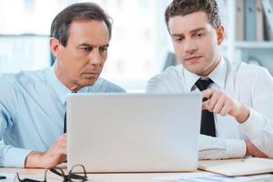 Searching for smart solutions together. Two serious business people in formalwear discussing something and looking at laptop while sitting at working place photo
