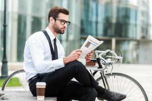 leyendo las últimas noticias. vista lateral de un joven hombre de negocios leyendo el periódico mientras se sienta cerca de su bicicleta con un edificio de oficinas al fondo foto