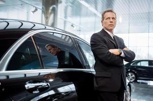 Dreaming of a new car. Thoughtful grey hair man in formalwear leaning at the car and looking away photo