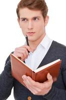 He got creative mind. Thoughtful young man holding note pad and touching chin with pen while standing isolated on white photo