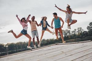 joven y lleno de energía. toda la longitud de los jóvenes con ropa informal sonriendo y gesticulando mientras saltan en el muelle foto