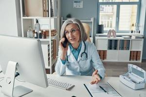 Confident mature female doctor in white lab coat talking on the phone and smiling while sitting in her office photo