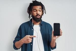 Charming young African man in casual wear pointing copy space and listening music while standing against grey background photo