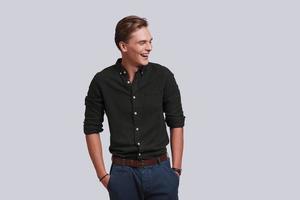 Used to look perfect. Good looking young man keeping hands in pockets and laughing while standing against grey background photo