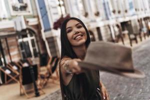 chica de moda mujer joven atractiva lanzando un sombrero y sonriendo mientras está de pie al aire libre foto