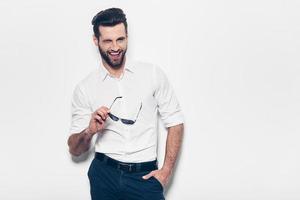 coqueto guapo. un joven apuesto con camisa blanca sosteniendo gafas de sol y guiñándote un ojo con una sonrisa mientras se enfrenta a un fondo blanco foto