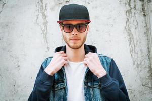 Confidence is a key to success. Handsome young man in headwear adjusting his jacket while standing against the concrete wall photo