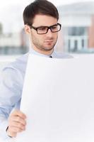 Confident architect. Confident young man in shirt and tie examining blueprint while standing against the window photo