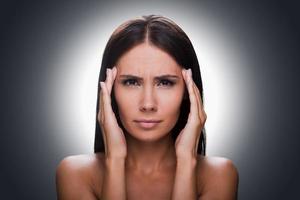 Terrible headache. Portrait of frustrated young shirtless woman looking at camera and touching head with hands while standing against grey background photo