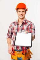 I need your signature here. Cheerful young handyman in hardhat holding stretching out clipboard with paper and smiling while standing against grey background photo