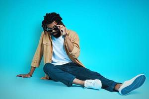 Handsome young African man in casual wear smiling and adjusting eyewear  while sitting against blue background photo