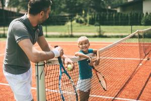 listo para jugar alegre padre e hija apoyándose en la red de tenis y mirándose con una sonrisa foto