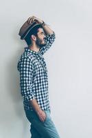 Stylish handsome. Side view of handsome young cheerful man holding one hand in pocket and adjusting his hat photo