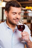 disfrutando del vino en el restaurante. apuesto joven sosteniendo un vaso con vino tinto y oliéndolo mientras está sentado en el restaurante foto