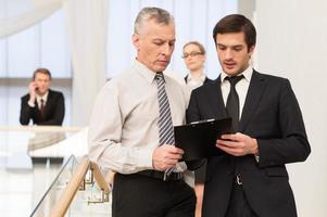 Asking an advice. Two men in formalwear discussing something while moving by staircase with people on background photo