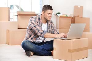 Working in brand new house. Concentrated young man sitting on the floor and working on laptop while cardboard boxes laying on the background photo