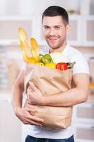 He is passionate about healthy food. Happy young man holding shopping bag full of groceries while standing in the kitchen photo