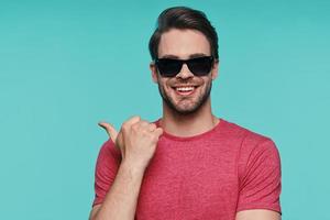 Portrait of handsome young man in sunglasses looking at camera and pointing copy space photo