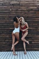 Young and full of energy. Full length of two attractive young women in swimwear laughing while standing against the wooden wall outdoors photo