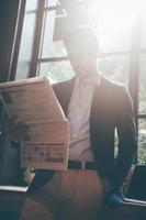 Reading fresh newspaper. Low angle view of confident young man reading fresh newspaper while leaning at the window sill in office or cafe photo