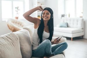 Enjoying best song ever Attractive young woman in headphones holding smart phone and gesturing while sitting on the couch at home photo