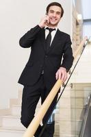 Always in touch. Cheerful young man in formalwear talking on the mobile phone while moving down by staircase photo