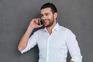 Good business talk. Handsome young man talking on the mobile phone and smiling while standing against grey background photo