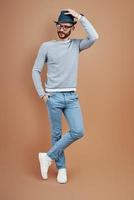 Full length of fashionable young man in casual wear adjusting hat while standing against brown background photo