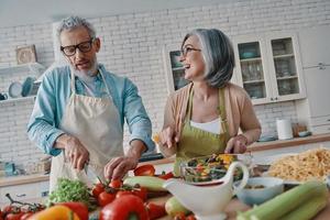 feliz pareja mayor en delantales preparando una cena saludable y sonriendo mientras pasa tiempo en casa foto