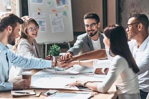 Teamwork. Young modern people in smart casual wear holding hands on top of one another in a symbol of unity while working in the creative office photo