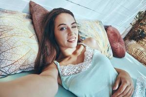 Beautiful young woman making selfie and smiling while lying down in bed photo