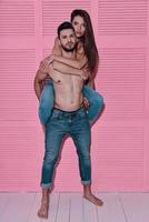 She means everything to him. Full length of handsome shirtless man carrying his girlfriend on shoulders while standing against pink background photo