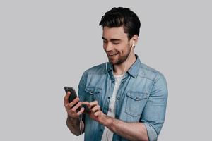 disfrutando de su música favorita. un joven apuesto con camisa de jeans azules escuchando música usando su teléfono inteligente y sonriendo mientras se enfrenta a un fondo gris foto