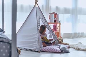 Enjoying her little world. Lovely little girl reading a book and smiling while sitting on the floor in tent photo