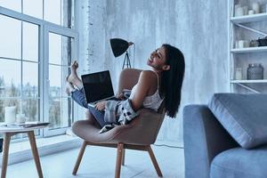 Enjoying every minute. Attractive young smiling woman looking away and using her laptop while sitting in armchair at home photo