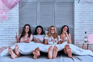 amigos para siempre. cuatro atractivas mujeres jóvenes en pijama bebiendo cócteles y sonriendo mientras están en la cama foto